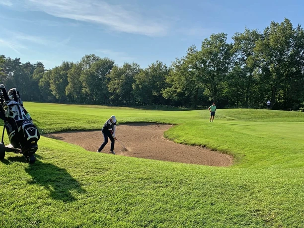 Ein Golfer spielt aus einem Sandbunker auf einem grünen Golfplatz, umgeben von Bäumen.