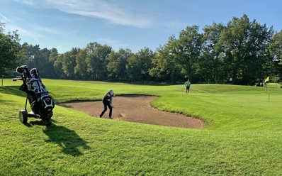 Golfspielen_GolfclubPeckeloh.jpeg Ein Golfer spielt aus einem Sandbunker auf einem grünen Golfplatz, umgeben von Bäumen.