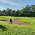 Golfspielen_GolfclubPeckeloh.jpeg Ein Golfer spielt aus einem Sandbunker auf einem grünen Golfplatz, umgeben von Bäumen.