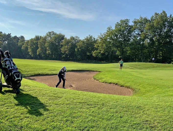 Golfspielen_GolfclubPeckeloh.jpeg Ein Golfer spielt aus einem Sandbunker auf einem grünen Golfplatz, umgeben von Bäumen.