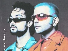 Zwei Männer mit Sonnenbrillen, einer mit Bart und Locken, der andere mit glattem, blau gefärbtem Haar.