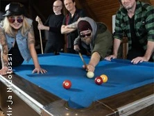 Fünf Personen spielen Billard in einem rustikalen Raum, mit konzentrierten Positionen am Tisch.