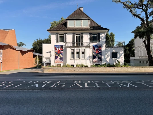 Das Bild zeigt ein weißes Haus mit braunem Dach, flankiert von zwei farbigen Kunstplakaten.