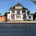 Ahlers Pro Arte_SAPA_Goebenstrasse.jpg Das Bild zeigt ein weißes Haus mit braunem Dach, flankiert von zwei farbigen Kunstplakaten.