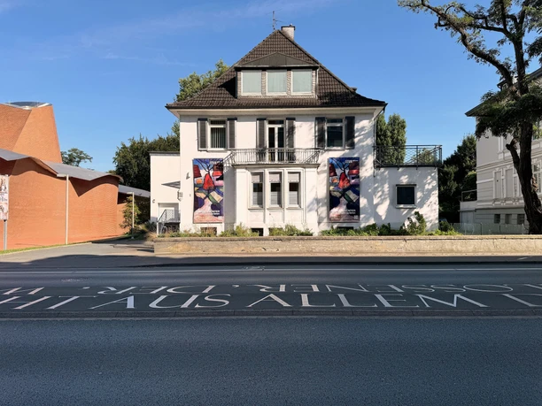 Ahlers Pro Arte_SAPA_Goebenstrasse.jpg Das Bild zeigt ein weißes Haus mit braunem Dach, flankiert von zwei farbigen Kunstplakaten.