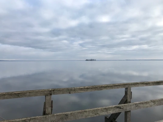 Vogelbeobachtungsturm Winzlar am Steinhuder Meer Holzgeländer vor stiller Gewässerlandschaft mit bewölktem Himmel, im Hintergrund eine kleine Insel.