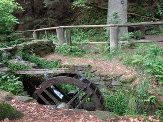 Mühle im Eifgenbachtal Altes hölzernes Wasserrad im Grünen, umgeben von Bäumen und einem rustikalen Holzzaun.