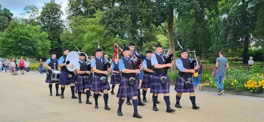 Eine Gruppe von Dudelsackspielern in schottischen Kilts marschiert durch einen Parkweg, umgeben von Bäumen.