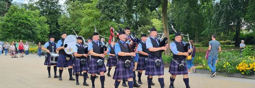 20240817_143612-74b1e5b7-6e4e3d30@800w.jpg Eine Gruppe von Dudelsackspielern in schottischen Kilts marschiert durch einen Parkweg, umgeben von Bäumen.
