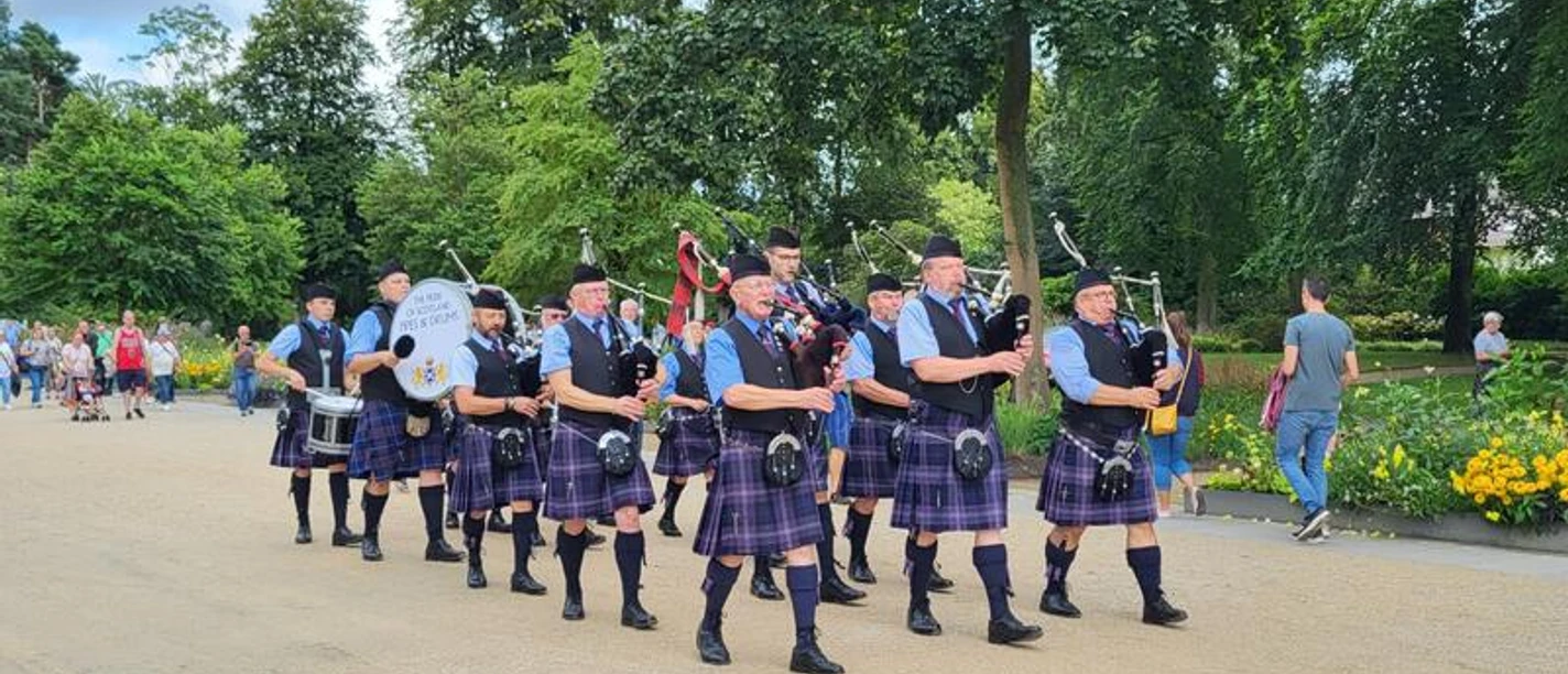 20240817_143612-74b1e5b7-6e4e3d30@800w.jpg Eine Gruppe von Dudelsackspielern in schottischen Kilts marschiert durch einen Parkweg, umgeben von Bäumen.