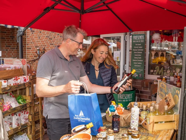 Shopping-in-Bad-Zwischenahn-Spezialitaeten.jpg Ein Paar beim Einkaufen lokaler Spezialitäten auf einem Marktstand in Bad Zwischenahn. Sie lächeln und halten Produkte in den Händen.
