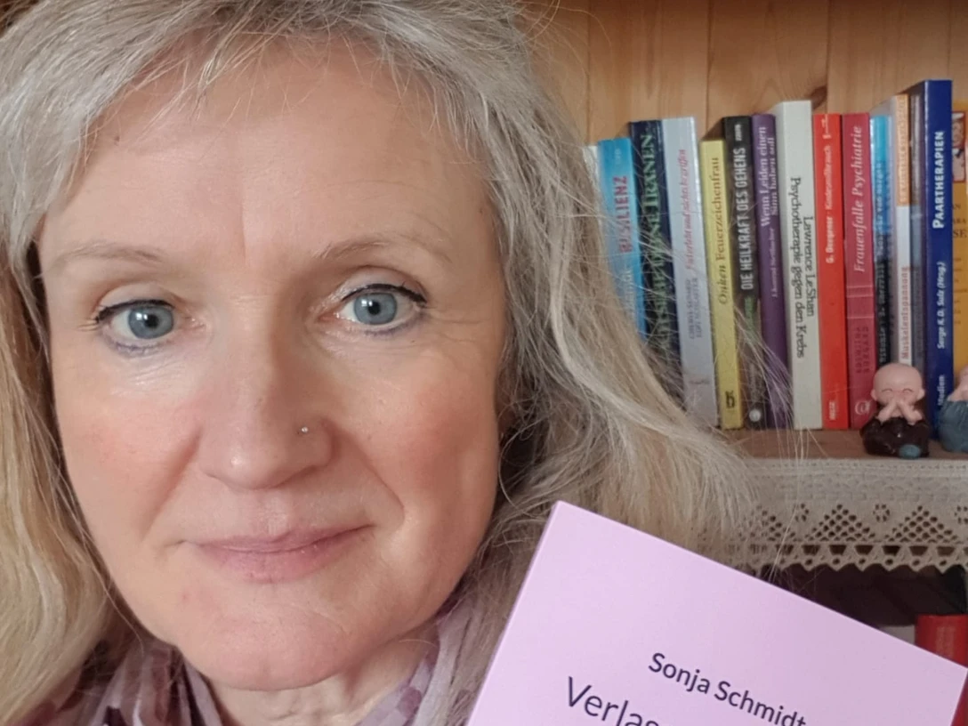Sonja Schmidt Sonja Schmidt trägt eine Bluse und hält ihr Buch "Verlassene Mütter, verlassene Töchter" vor einem Bücherregal.