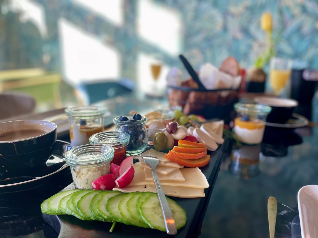 Frühstückstisch mit Kaffee, Käse, Gurken und Aufstrichen, Körbchen mit Brötchen im Hintergrund