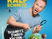 Ralf Schmitz in einer dynamischen Pose mit Lupe vor einem blauen Hintergrund und verschiedenen Requisiten.