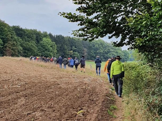 Whisky Wanderung Eine Wandergruppe spaziert am Rand eines frisch gepflügten Feldes entlang, flankiert von Bäumen.