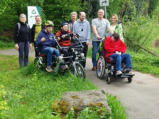 Inklusion Eine Gruppe von acht Personen, davon drei im Rollstuhl, genießt einen Spaziergang auf einem Waldweg.