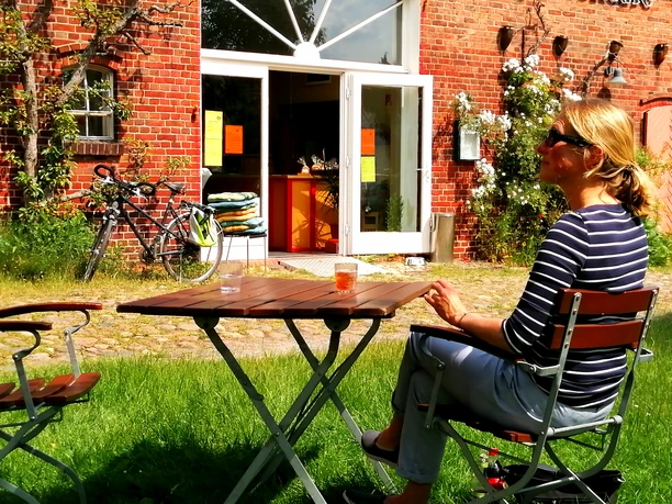 Mühlencafé von Rönn Eine Frau sitzt an einem Holztisch auf der Terrasse vor dem Eingang des Mühlencafé.