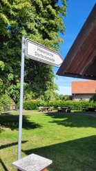 Hille Holzhausen II - Alte Scheune und Historische Dorfschmiede Hinweisschild zur Historischen Dorfschmiede neben einem grünem Bereich mit Bänken und Bäumen.