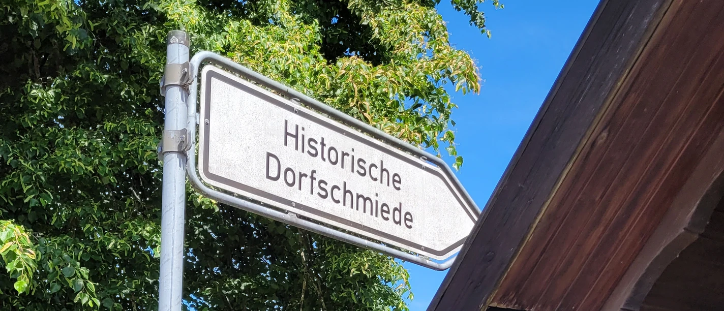 Hille Holzhausen II - Alte Scheune und Historische Dorfschmiede Hinweisschild zur Historischen Dorfschmiede neben einem grünem Bereich mit Bänken und Bäumen.