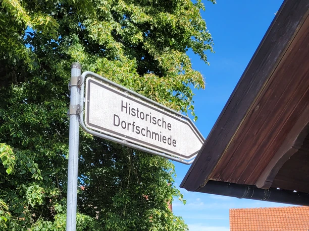 Hille Holzhausen II - Alte Scheune und Historische Dorfschmiede Hinweisschild zur Historischen Dorfschmiede neben einem grünem Bereich mit Bänken und Bäumen.