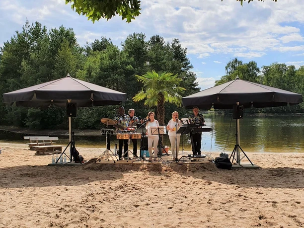 Foto 2 Sal Y Mango.jpg Een band speelt onder parasols op het zandstrand, omringd door bomen en een rustig meer.