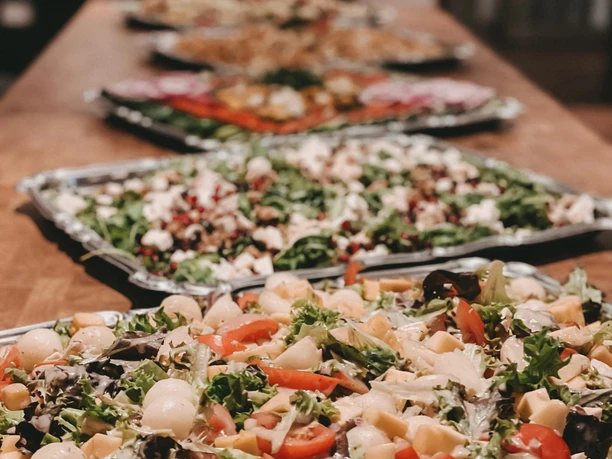 Verschiedene reichhaltig belegte Salatplatten stehen auf einem langen Holztisch, Hintergrund unscharf.
