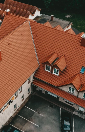 Luftbild eines großen Gebäudekomplexes mit rotem Ziegeldach, umgeben von parkenden Autos und Bäumen.