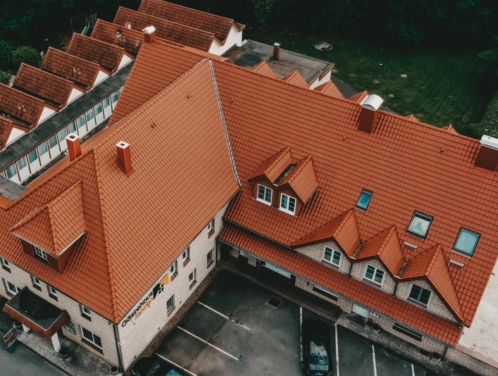Luftbild eines großen Gebäudekomplexes mit rotem Ziegeldach, umgeben von parkenden Autos und Bäumen.