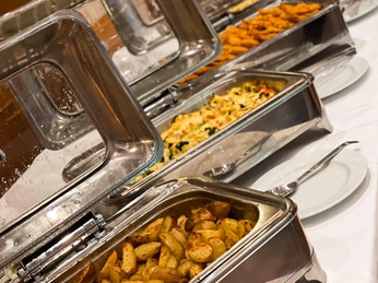 Verschiedene Speisen in Edelstahlschalen: Kartoffelspalten, Gemüseauflauf und paniertes Fleisch.