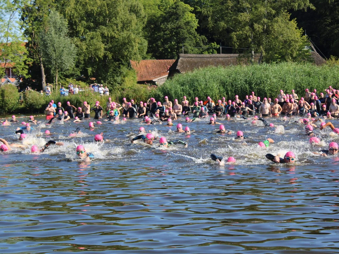 quer-durchs-meer-schwimmerfeld2.jpg Inmitten eines Sees schwimmen viele Menschen mit pinken Badekappen im Wasser; dicht gedrängt sind sie von üppigem Grün und zahlreichen Zuschauern am Ufer umgeben.