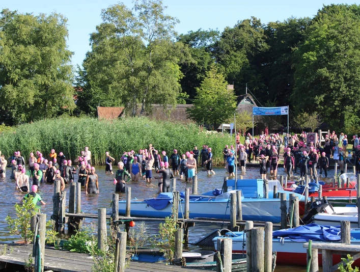 quer-durchs-meer-start-dreibergen.jpg Ein spannendes Schwimmevent am Zwischenahner Meer in Bad Zwischenahn, mit zahlreichen Teilnehmern in bunten Badehauben und eine umringende grüne Naturkulisse.