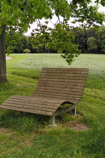 Waldsofa an der Wilhelmshöhe Stemwede Holzliegebank unter einem Baum an einem Wanderweg, mit Blick auf grüne Wiesen und Wälder.