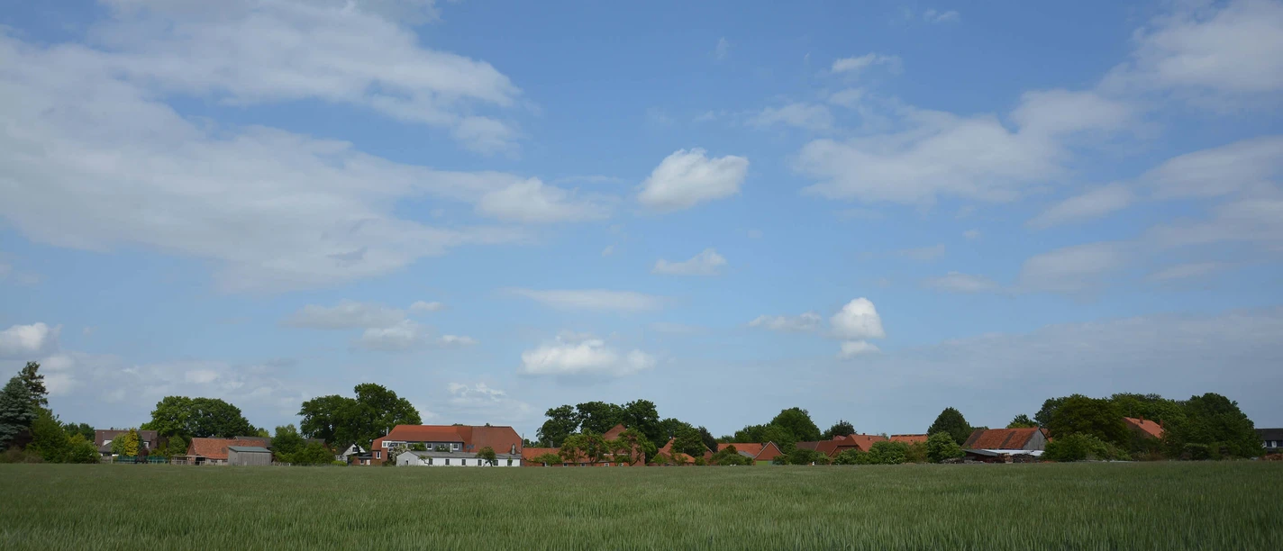 suttorf Eine weitläufige grüne Wiese erstreckt sich vor einer kleinen Ansammlung von Backsteinhäusern unter blauem Himmel.