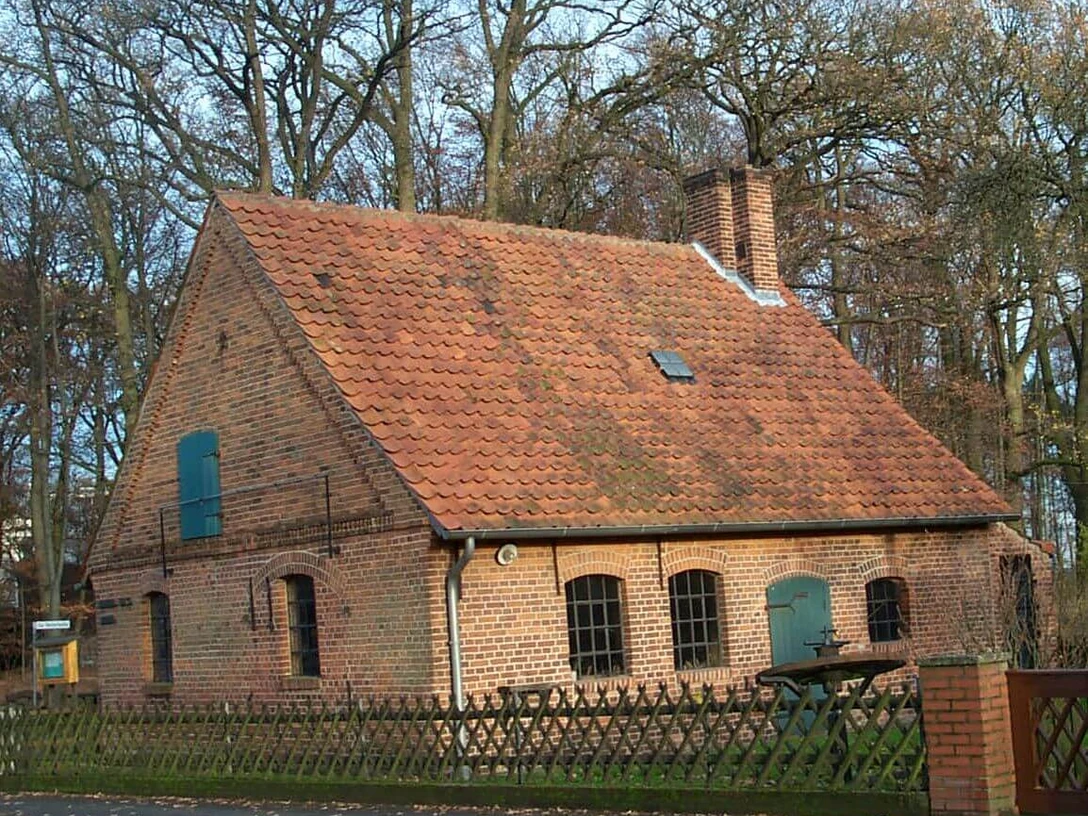 helstorf-schmiede Eine historische Schmiede aus Backstein mit roten Dachziegeln, umgeben von einem Holzzaun und Bäumen.