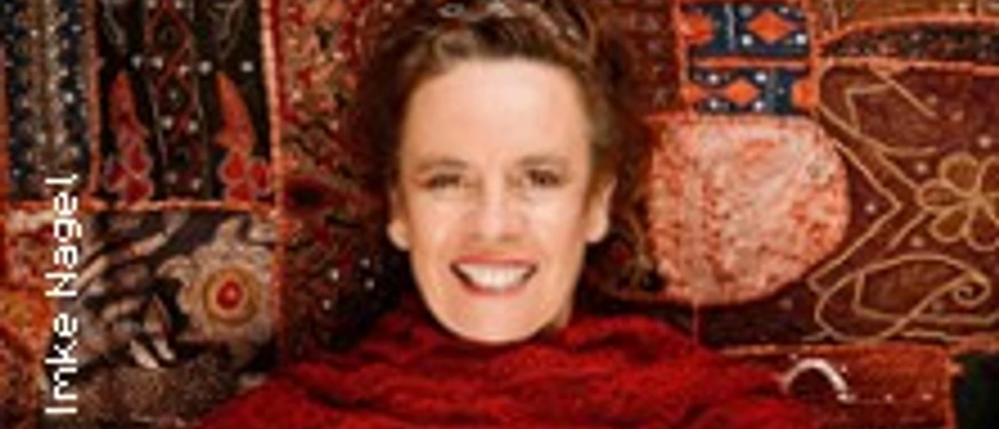 Eine lächelnde Frau in einem leuchtend roten Schal vor einem reichen, detailreichen Wandteppich.