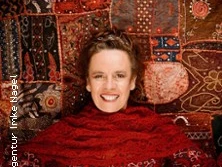 Eine lächelnde Frau in einem leuchtend roten Schal vor einem reichen, detailreichen Wandteppich.