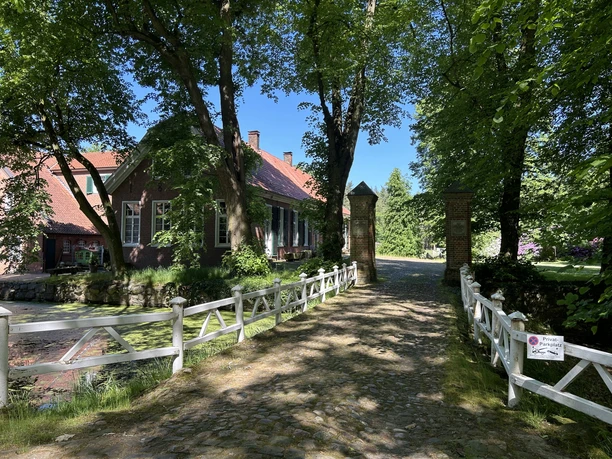 Gut Düneburg, Haren (Ems) – Außenansicht Herrenhaus Backsteingebäude mit rotem Dach hinter schattiger Allee und weißem Zaun an einem Wassergraben.