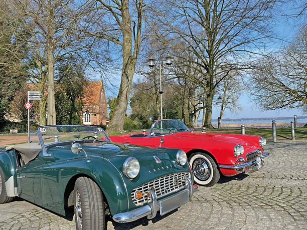 Classic-Car-Charity-Bad-Zwischenahn4.jpg Zwei klassische Sportwagen, ein grüner Triumph TR2 und ein roter Chevrolet Corvette C1, parken auf einer gepflasterten Fläche unter kahlen Bäumen im Sonnenschein.