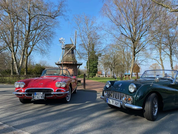 Classic-Car-Charity-Bad-Zwischenahn1.jpg Ein roter Chevrolet Corvette und ein grüner Triumph TR3 sind vor der historischen Windmühle mit blauem Himmel im Hintergrund zu sehen.