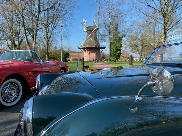 Classic-Car-Charity-Bad-Zwischenahn2.jpg Ein klassisches Auto glänzt im Vordergrund, während im Hintergrund eine historische Mühle steht. Bäume rahmen die Szene ein, ein Hauch von Nostalgie in Bad Zwischenahn.