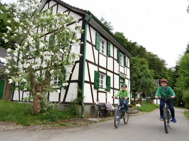 Dhünn-Runde Radfahrer vor einem traditionellen Fachwerkhaus auf einer ländlichen Straße im Grünen.