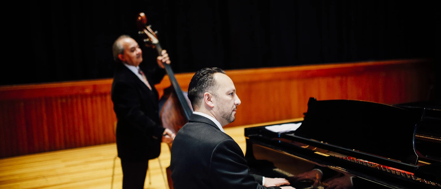 Ein Pianist spielt an einem Klavier, während ein Mann im Hintergrund einen Kontrabass hält. Beide sind elegant gekleidet und befinden sich auf einer Bühne.
