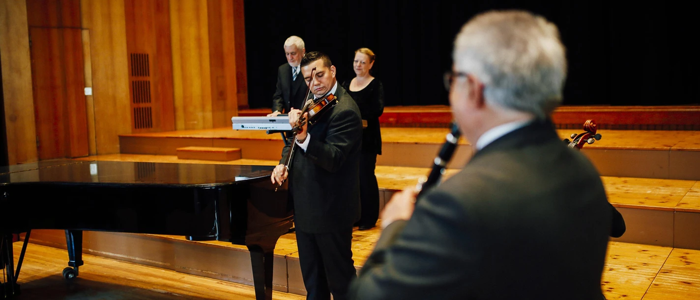 Eine musikalische Aufführung mit einem Violinisten und einem Bläser auf einer Bühne. Im Hintergrund sind weitere Musiker zu sehen, die sich bereit machen.
