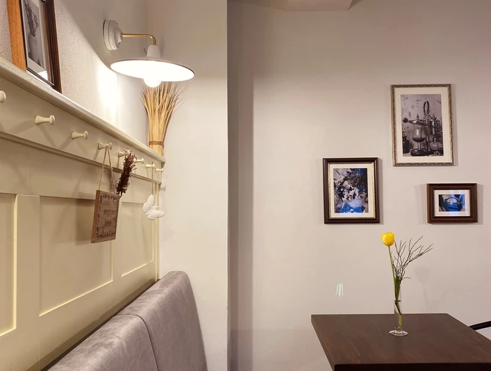 Münte1.jpg Gemütliche Café-Ecke mit grauer Sitzbank, Holztisch, gelber Blume und gerahmten Bildern an der Wand.