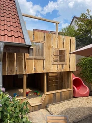 Münte2.jpg Holzspielhaus mit roter Rutsche und Sandkasten in kleinem Garten, umgeben von Pflanzen und einem Haus.