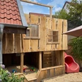 Münte2.jpg Holzspielhaus mit roter Rutsche und Sandkasten in kleinem Garten, umgeben von Pflanzen und einem Haus.