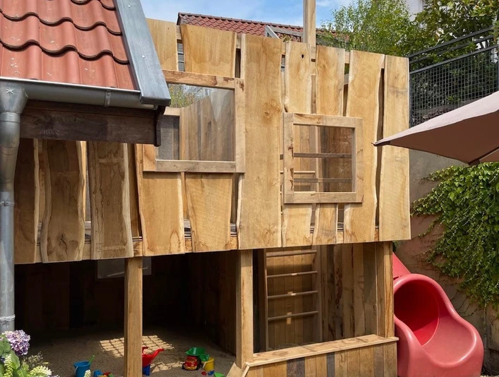 Münte2.jpg Holzspielhaus mit roter Rutsche und Sandkasten in kleinem Garten, umgeben von Pflanzen und einem Haus.