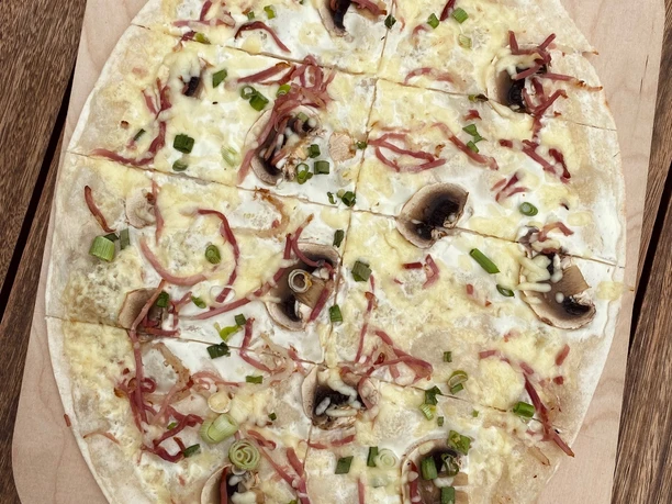 Ein knuspriger Flammkuchen auf Holzbrett mit Pilzen, Zwiebeln und Speck, in acht Stücke geschnitten.