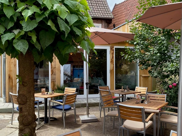 Münte7.jpg Gemütlicher Außenbereich eines Cafés mit Holzstühlen, Tischdekoration und schattenspendenden Schirmen.