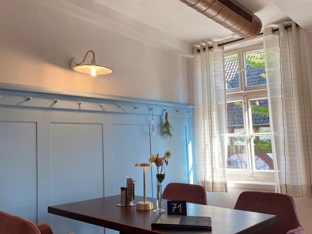 Münte8.jpg Ein gemütlicher Café-Tisch mit vier rosa Stühlen in einem hellen Raum mit Fenster und dekorativer Wand.